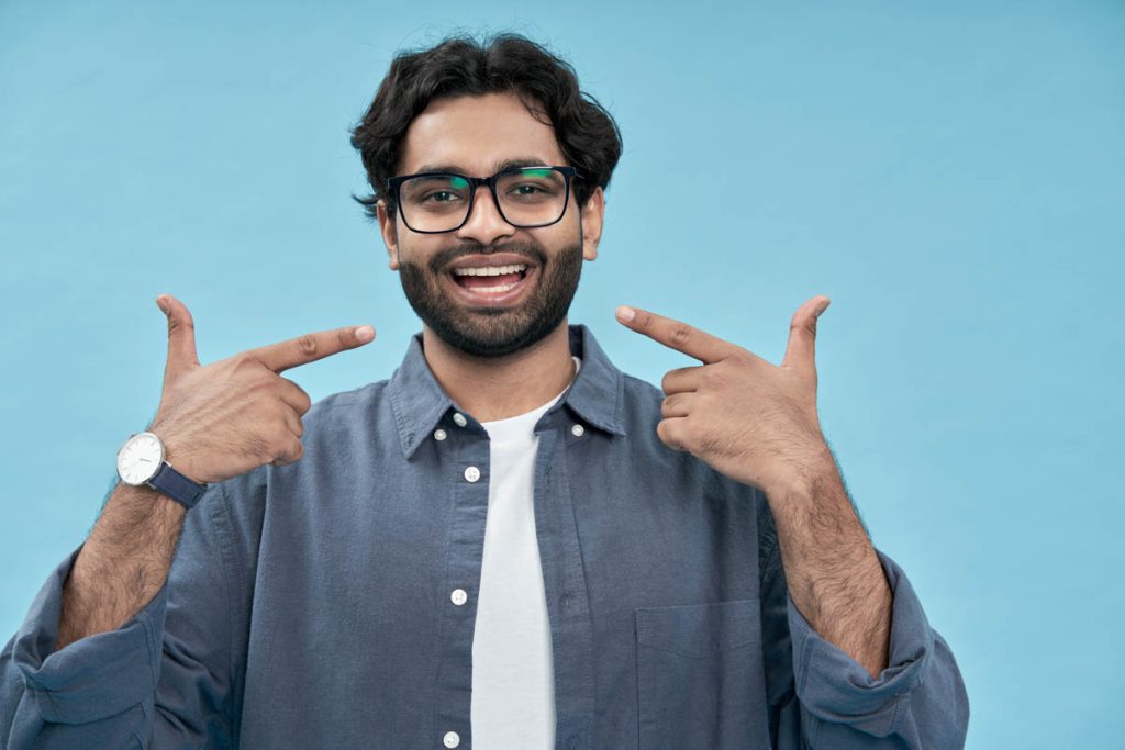 Happy man pointing fingers at healthy white teeth advertising dentist services or dental care product commercial promo, presenting perfect smile standing isolated on blue background.